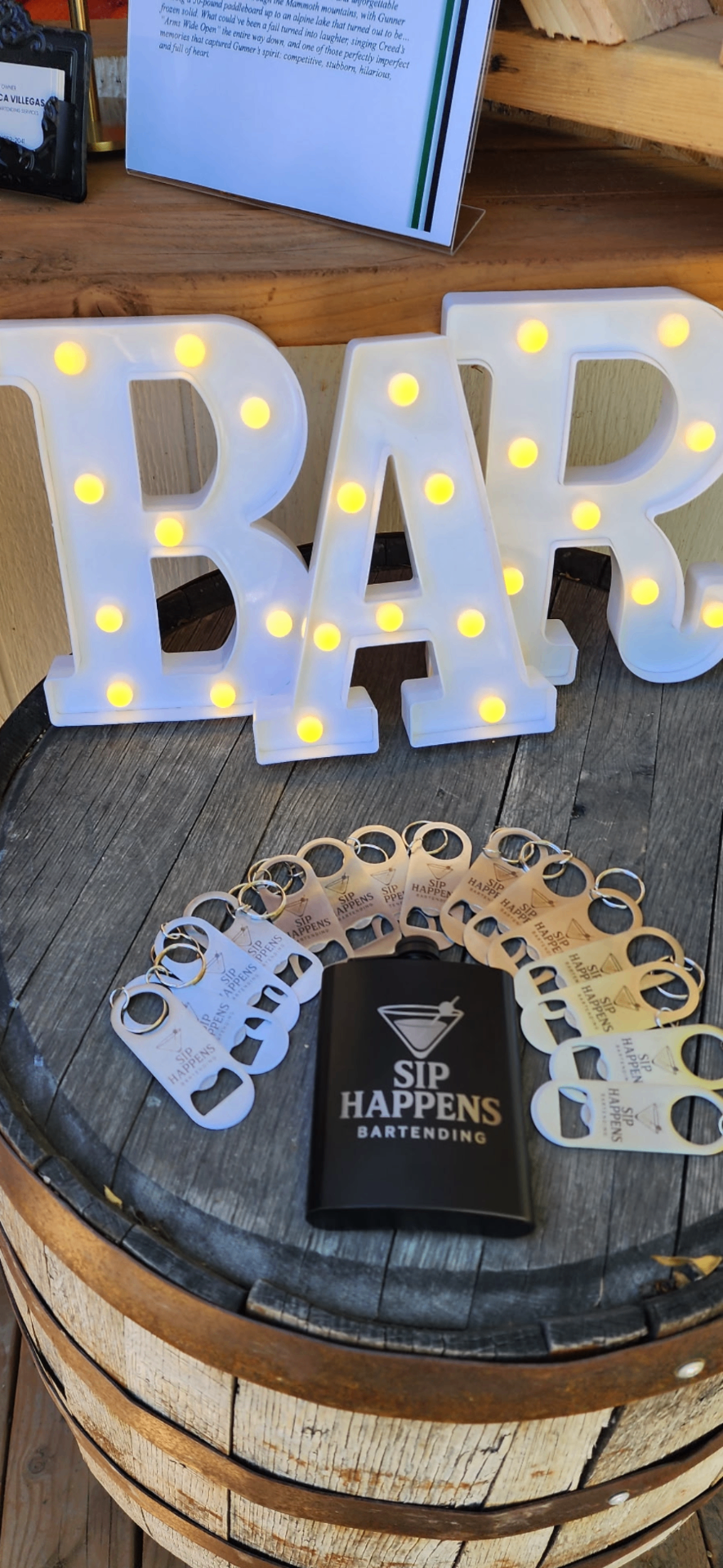 Glowing 'BAR' marquee sign on a rustic barrel with the Sip Happens Bartending flask and keychains on display