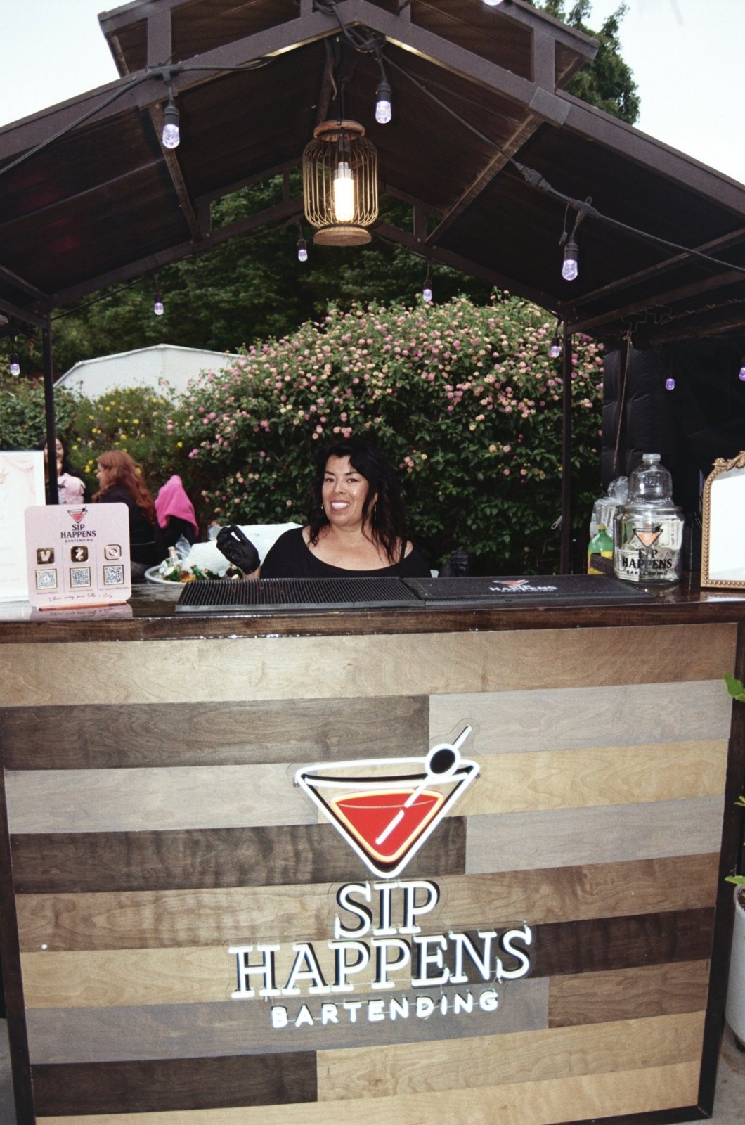 Rustic wood Sip Happens Bartending bar with a neon sign under a lit patio structure at a garden event